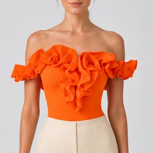 Puede incluir: Un top naranja vibrante con hombros descubiertos que presenta un diseño de volantes superpuestos prominente en el pecho y los hombros, combinado con pantalones de color beige claro. El top tiene un corpiño ajustado y escote recto.
