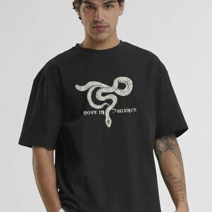 以下が含まれることがあります： 白いヘビのデザインと「MOVE IN SILENCE」の文字が入った黒いTシャツ。ヘビの頭には緑色の宝石が付いています。このシャツはクルーネックで半袖です。