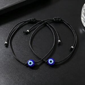 Puede incluir: Dos pulseras de cordón negro ajustables, cada una con una cuenta circular azul de ojo turco. Las pulseras cuentan con cierres de nudo corredizo y pequeñas cuentas de metal plateado en los extremos de los cordones, con un diseño sencillo.
