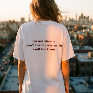 May include: A white short-sleeved t-shirt featuring dark brown text printed on the back that reads: i'm not rihanna, i don't love the way you lie, i will block you. The garment has a crew neckline and a standard fit, shown from the back view.