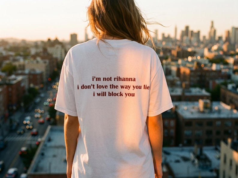 May include: A white short-sleeved t-shirt featuring dark brown text printed on the back that reads: i'm not rihanna, i don't love the way you lie, i will block you. The garment has a crew neckline and a standard fit, shown from the back view.