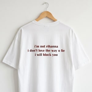 May include: A white short-sleeved t-shirt featuring dark red text printed on the back. The text reads: i'm not rihanna, i don't love the way u lie, i will block you. The shirt is displayed on a wooden hanger against a plain white background.