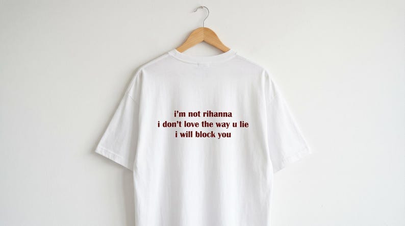May include: A white short-sleeved t-shirt featuring dark red text printed on the back. The text reads: i'm not rihanna, i don't love the way u lie, i will block you. The shirt is displayed on a wooden hanger against a plain white background.