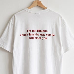 May include: A white short-sleeved t-shirt featuring red text printed on the back that reads: i'm not rihanna, i don't love the way you lie, i will block you. The shirt is displayed on a wooden hanger against a plain white background.
