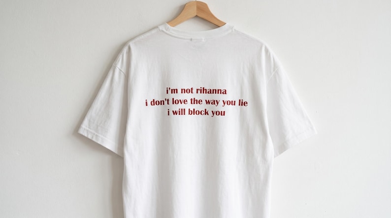 May include: A white short-sleeved t-shirt featuring red text printed on the back that reads: i'm not rihanna, i don't love the way you lie, i will block you. The shirt is displayed on a wooden hanger against a plain white background.