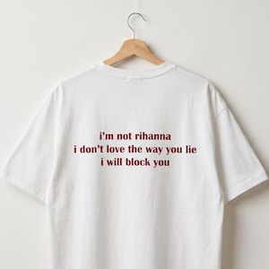 May include: White t-shirt with burgundy text: "I'm not rihanna, I don't love the way you lie, I will block you". The tee is suspended on a wooden hanger against a white backdrop.