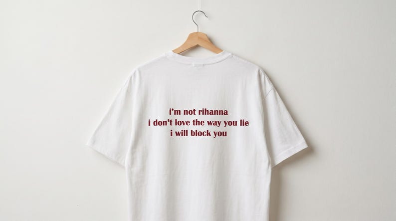 May include: A white short-sleeved t-shirt hanging on a wooden hanger. The back of the shirt features dark red text that reads: i'm not rihanna, i don't love the way you lie, i will block you. The garment has a crew neckline and a plain white finish.