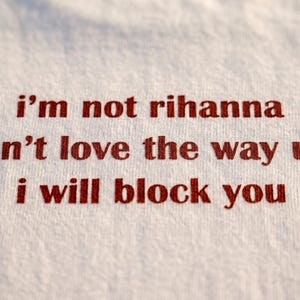 May include: A white fabric surface featuring red embroidered text that reads: i'm not rihanna, i don't love the way u lie, i will block you. The lettering is centred and uses a simple, lowercase font style against the textured white textile background.