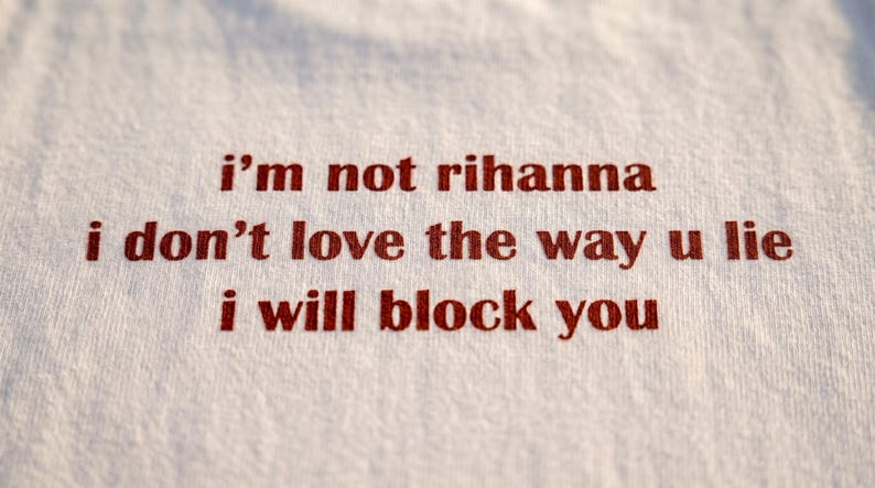 May include: A white fabric surface featuring red embroidered text that reads: i'm not rihanna, i don't love the way u lie, i will block you. The lettering is centred and uses a simple, lowercase font style against the textured white textile background.