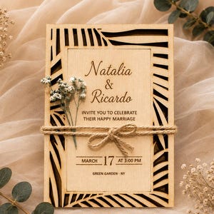 May include: A rectangular wooden wedding invitation card featuring a laser-cut palm leaf border. The center is engraved with the names Natalia & Ricardo, event details, and the date March 17 at 3:00 PM. A jute twine bow secures a sprig of baby's breath.