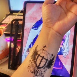Puede incluir: Tatuaje temporal negro aplicado en el antebrazo. Presenta un diseño de escudo con alas, elementos florales y un borde en forma de engranaje. El diseño es detallado e intrincado, con líneas finas y sombreado.