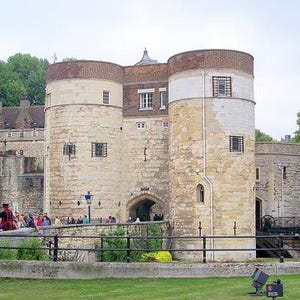 Könnte beinhalten: Außenansicht des Tower of London, einer historischen Burg mit Steinmauern und zylindrischen Türmen. Die Türme haben Ziegel- und weiße Akzente. Eine Gruppe von Menschen versammelt sich in der Nähe des Eingangs, mit grünen Bäumen im Hintergrund.
