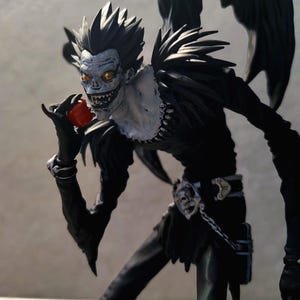 May include: A detailed figurine of a tall, slender, humanoid creature with spiky black hair, pale white skin, and glowing yellow eyes. The figure wears a black outfit with a silver belt buckle and holds a small, shiny red apple in its right hand.