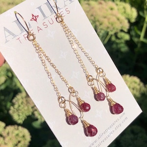 Könnte beinhalten: Goldene Ohrringe mit drei Tropfen aus rubinroten Edelsteinen an jedem Ohrring. Die Ohrringe befinden sich auf einer weißen Karte mit dem aufgedruckten Text "Alira Treasures".