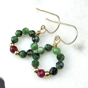 May include: A pair of hoop earrings featuring faceted green and red gemstone beads interspersed with small, polished gold-tone metal spheres. The beads are arranged in a circular pattern, suspended from simple gold-tone metal ear wires.