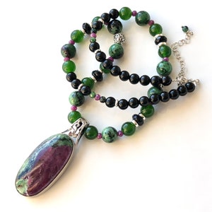 Peut inclure: Un collier avec un pendentif en pierre précieuse verte et rose avec des perles noires. Le pendentif est de forme ovale et a un sertissage argenté. Le collier est fait de perles vertes et noires.
