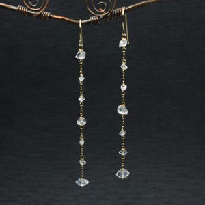 Puede incluir: Un par de aretes largos de cadena dorada con gotas de cristal transparente facetado. Los aretes están colgados de un soporte de alambre.