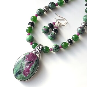 May include: A green and pink gemstone necklace and earrings set with a silver pendant and black accents.