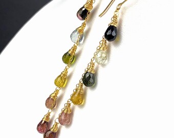 May include: Gold-toned dangle earrings featuring a cascade of colorful, teardrop-shaped gemstones. The gemstones include shades of pink, yellow, green, blue, and black. The earrings are suspended from delicate gold wires, creating a graceful and elegant design.