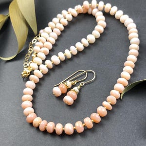 May include: A necklace and earring set featuring peach-colored beads and gold-colored accents. The necklace is composed of small, rounded beads strung together, with a matching pair of earrings. The jewelry set is displayed on a dark surface.