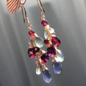 Könnte beinhalten: Hängeohrringe mit goldfarbenen Haken und Draht. Die Ohrringe zeigen Cluster aus facettierten Edelsteinen in den Farbtönen Tiefrot, Blau und Hellgrün. Die Edelsteine sind mit Draht umwickelt, wodurch ein zartes und elegantes Design entsteht.