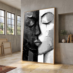 Puede incluir: Una gran obra de arte en lienzo enmarcada que representa un rostro dividido en blanco y negro. Un lado es negro, el otro blanco, con los ojos cerrados y una lágrima. La obra tiene un estilo texturizado, de impasto, en una habitación con luz natural.