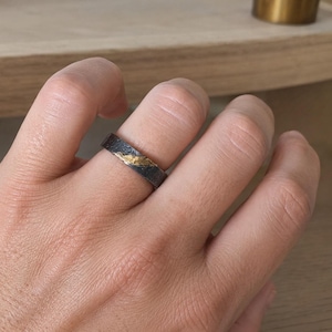 Può includere: Un anello scuro e strutturato con un intarsio color oro. L'anello è indossato su un dito, mostrando il suo design unico e i colori contrastanti. La fascia sembra essere fatta di un metallo scuro, possibilmente argento ossidato o un materiale simile.
