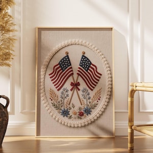 May include: Embroidered artwork featuring two American flags crossed at the base, with red and white stripes and a field of stars. The flags are set against a beige background, surrounded by a floral design and a cream-colored, beaded border, all within a gold frame.