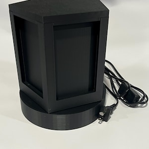 May include: A black, geometric lamp with a hexagonal top and a rectangular frame. The lamp sits on a circular base and has a power cord with a two-prong plug. The lamp is made of a matte black material.