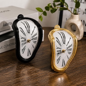 Peut inclure: Deux horloges analogiques au style fondu, l'une avec un cadre noir et l'autre avec un cadre doré. Toutes deux présentent des cadrans blancs avec des chiffres romains noirs et des aiguilles centrales. Elles ont une forme asymétrique.