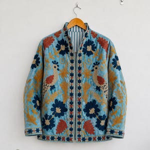 Pode incluir: Um casaco acolchoado azul-claro com bordados intrincados de flores azul-escuras, folhagem amarelo-dourada e dois pássaros com asas castanho-avermelhadas. O casaco tem gola mandarim, mangas compridas e uma abertura frontal com forro às riscas.