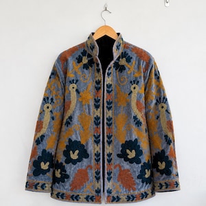 Puede incluir: Una chaqueta de terciopelo azul claro con intrincados bordados de aves estilizadas y grandes motivos florales en tonos azul oscuro, amarillo mostaza y naranja quemado. La chaqueta tiene cuello alto, mangas largas y diseño abierto.