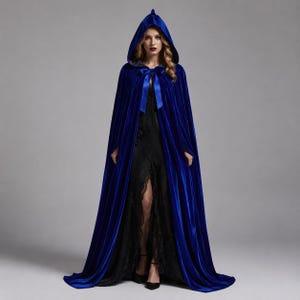May include: A floor-length royal blue velvet hooded cloak featuring a matching ribbon tie closure at the neck. The cape is worn over a black lace dress with a high front slit, showcasing a deep blue fabric with a soft, reflective sheen.