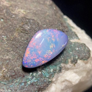 May include: A polished, teardrop-shaped opal gemstone featuring a translucent light blue base color with vibrant, multi-colored flashes of red, orange, and yellow light reflecting across its smooth, domed surface in a distinct play of color.
