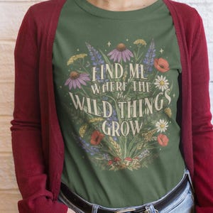 May include: Green t-shirt with the text "FIND ME WHERE THE WILD THINGS GROW" surrounded by colorful wildflowers. The person is wearing a burgundy cardigan and blue jeans.
