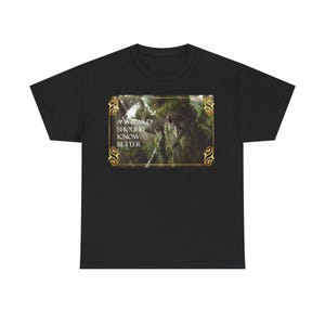 Op de afbeelding: Zwart T-shirt met een ingelijste afbeelding van een tovenaar in een bos. De afbeelding heeft de tekst "A WIZARD SHOULD KNOW BETTER" in het wit. De lijst heeft een gouden filigraan ontwerp.