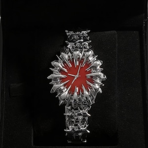 May include: A silver-toned wristwatch with a striking design, featuring a red watch face. The watch is presented in a black box, highlighting its intricate, spiky detailing. The watch band and bezel have a unique, textured appearance.