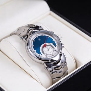 Pu&ograve; includere: Orologio da polso argentato con quadrante blu e lancetta dei secondi rossa, presentato in una scatola nera con interno bianco. L'orologio ha un design unico con un quadrante bianco pi&ugrave; piccolo e un cinturino argentato.