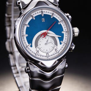 Pu&ograve; includere: Orologio da polso in acciaio inossidabile con quadrante blu e quadrante bianco pi&ugrave; piccolo. L'orologio ha una lancetta dei secondi rossa e un'indicazione della data alle 6. Il cinturino &egrave; anch'esso in acciaio inossidabile.