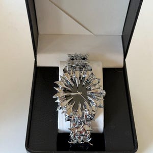 May include: A silver-toned wristwatch with a unique spiky design, presented in a black box with a white interior. The watch features a dark watch face and a matching spiky band. The watch is a statement piece.