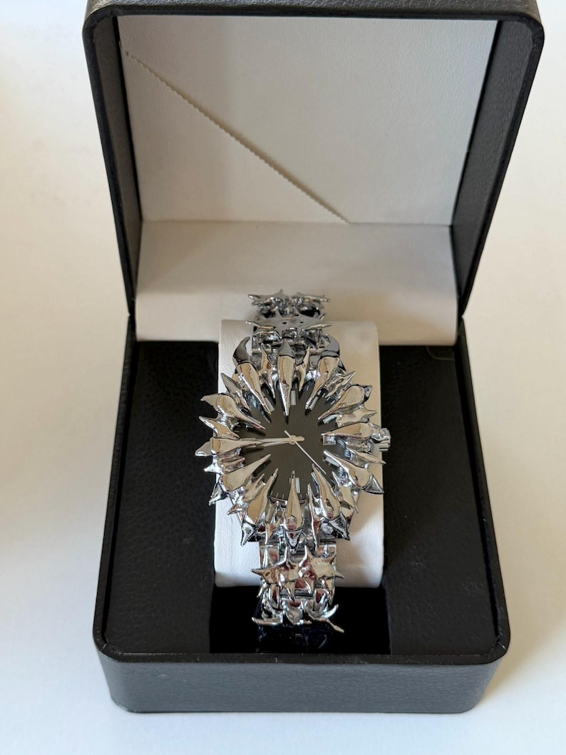 May include: A silver-toned wristwatch with a unique spiky design, presented in a black box with a white interior. The watch features a dark watch face and a matching spiky band. The watch is a statement piece.