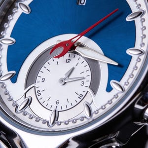 Pu&ograve; includere: Primo piano di un orologio da polso di lusso con quadrante blu e dettagli argentati. L'orologio presenta un quadrante secondario, un datario alle 6 e una lancetta dei secondi rossa. Il cinturino &egrave; in acciaio inossidabile.