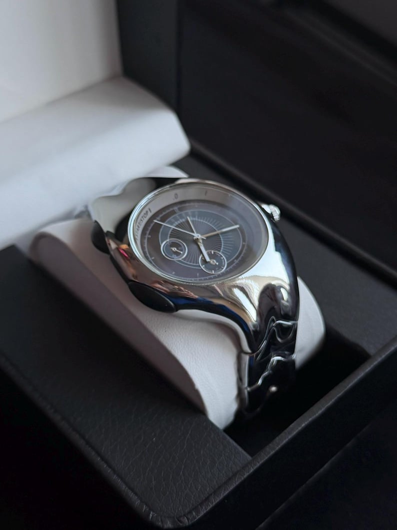 May include: A silver-toned wristwatch with a dark gray face and three sub-dials, presented in a black box with a white cushion. The watch has a unique, curved design and a matching silver band.