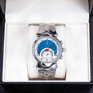 Pu&ograve; includere: Un orologio da polso argentato con quadrante blu e cinturino argentato &egrave; presentato in una scatola nera con interno bianco. L'orologio ha un quadrante rotondo con lancette e indicatori argentati. Il cinturino &egrave; composto da maglie argentate.