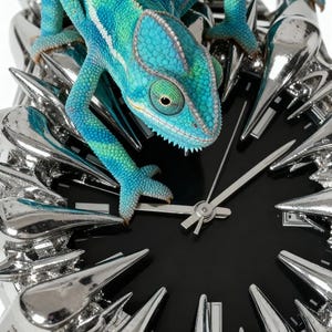 May include: A unique clock featuring a vibrant blue and green chameleon perched on the face. The clock has a black dial with silver hands and is surrounded by a spiky, metallic silver frame. The overall design is artistic and eye-catching.