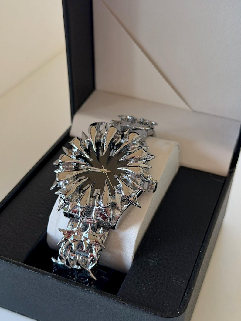 May include: A silver-toned wristwatch with a unique spiky design, presented in a black box with a white interior. The watch features a dark watch face and a textured band, creating a striking visual contrast.
