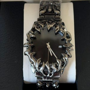 Puede incluir: Un reloj de pulsera negro con un dise&ntilde;o de bisel y correa ornamentado. La esfera del reloj es negra con manecillas plateadas y una pantalla de fecha. El reloj se presenta en una caja negra con interior blanco.