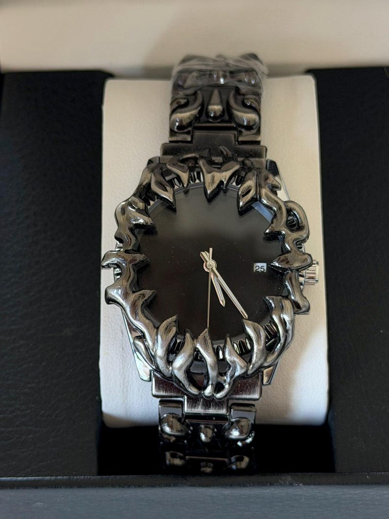 Puede incluir: Un reloj de pulsera negro con un dise&ntilde;o de bisel y correa ornamentado. La esfera del reloj es negra con manecillas plateadas y una pantalla de fecha. El reloj se presenta en una caja negra con interior blanco.