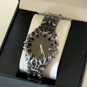 Puede incluir: Un reloj de pulsera negro con un dise&ntilde;o &uacute;nico en forma de llama alrededor de la esfera y en la correa. El reloj tiene una esfera negra con manecillas doradas y se presenta en una caja negra con un coj&iacute;n blanco.