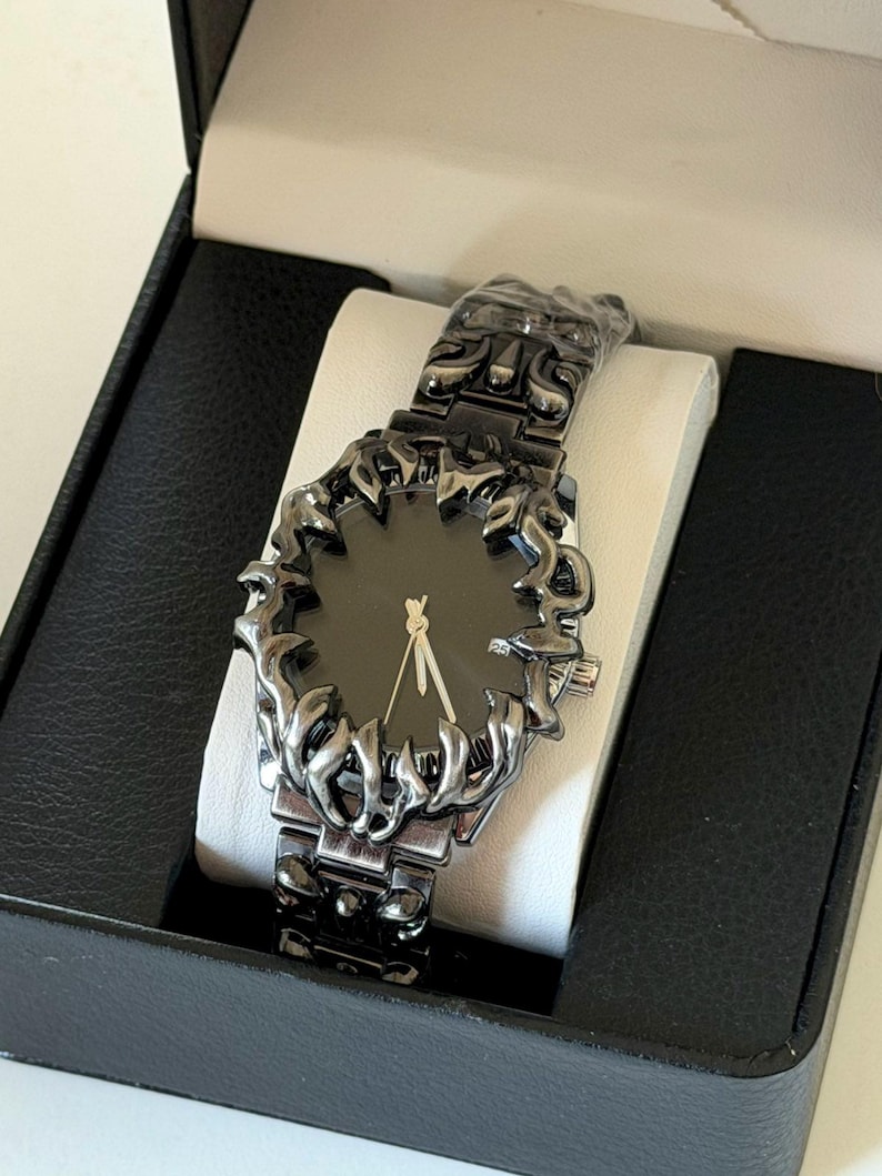 Puede incluir: Un reloj de pulsera negro con un dise&ntilde;o &uacute;nico en forma de llama alrededor de la esfera y en la correa. El reloj tiene una esfera negra con manecillas doradas y se presenta en una caja negra con un coj&iacute;n blanco.